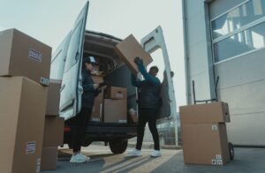 Workers loading boxes into a delivery van at a warehouse facility outdoors.